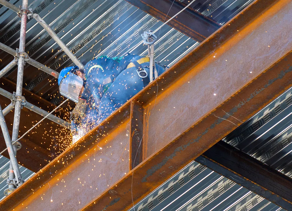 A welder in a visor and harness welding a steel beam on a building site, with sparks flying beside a scaffold